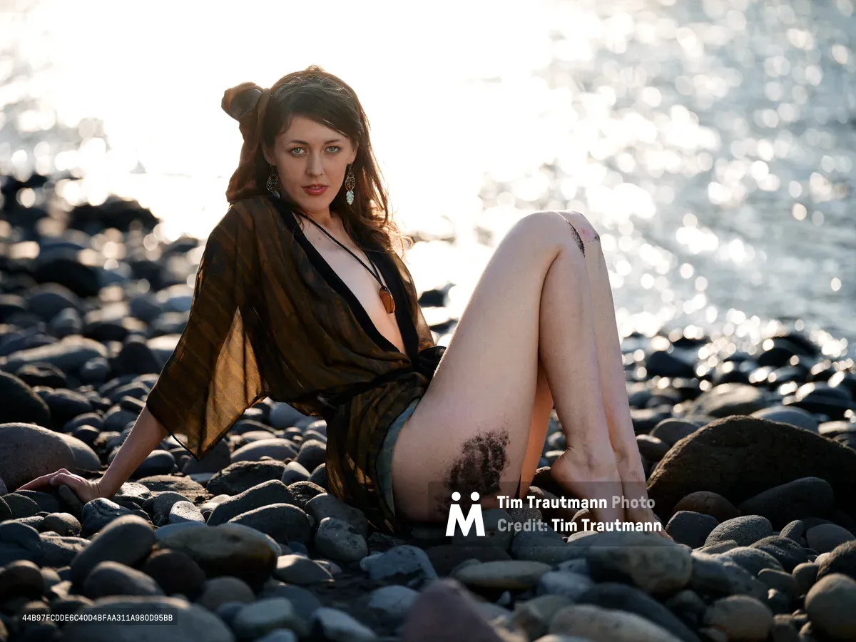 A woman with dark brown hair sits contemplatively among weathered river rocks along Oregon's Columbia River Gorge, her olive-toned wrap flowing around her figure. The late afternoon sun creates dramatic backlighting, casting her silhouette against sparkling water and transforming the rocky riverbed into a canvas of warm shadows and brilliant highlights. Her pendant necklace and dangling earrings catch the ethereal light as she gazes toward the camera with a serene, introspective expression.