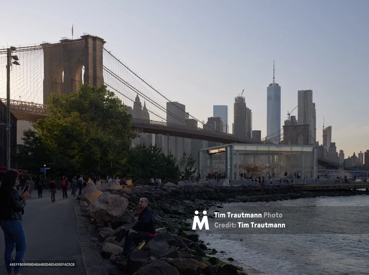 The iconic Brooklyn Bridge spans majestically across the East River as twilight settles over Lower Manhattan's gleaming skyline, including One World Trade Center. Visitors gather along the rocky shoreline of Brooklyn Bridge Park in DUMBO, some seated contemplatively on the weathered stones while others stroll the waterfront promenade. The modern glass pavilion of Jane's Carousel glows softly against the dusky sky, creating a serene juxtaposition between historic infrastructure and contemporary urban design.
