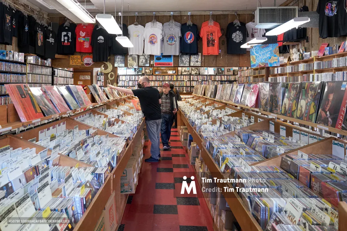 Two middle-aged men browse through extensive vinyl record collections during Record Store Day 2022 in the intimate aisles of Music Millennium on East Burnside Street. Fluorescent lighting illuminates towering wooden bins overflowing with albums while vintage band t-shirts dangle from the ceiling like musical totems. The red and black checkered floor creates a rhythmic pattern beneath their feet as they dig through decades of musical history in this beloved Portland institution during the annual celebration of independent record stores.