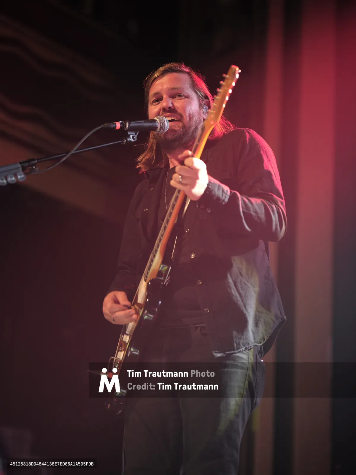 Russell Marsden, vocalist and guitarist of Band of Skulls, delivers an intimate performance at Webster Hall during the band's 10 Year Anniversary Tour. Bathed in warm amber stage lighting with deep red hues washing the background, Marsden leans into his microphone while gripping his electric guitar, his bearded face animated with the energy of live performance. The moody lighting creates dramatic shadows across the historic Manhattan venue, capturing the raw authenticity of this milestone celebration.