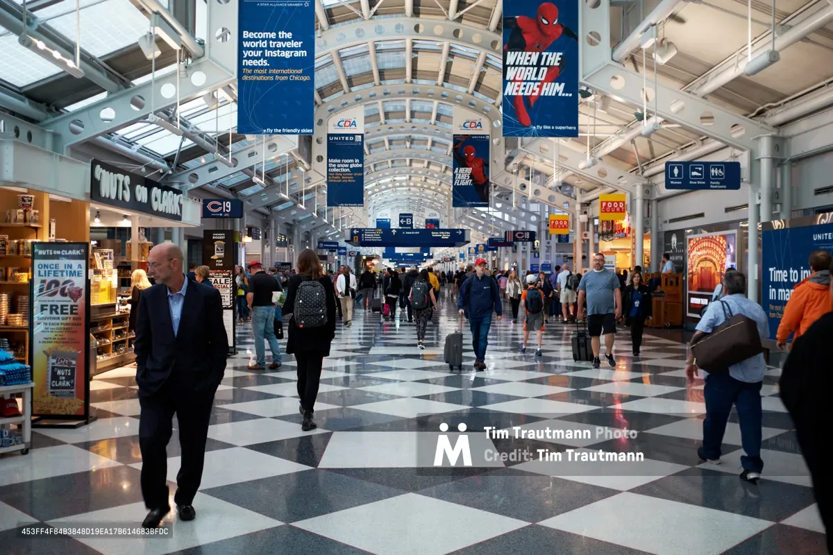 Travelers move through the bustling concourse of Chicago O'Hare International Airport, their silhouettes scattered across gleaming checkered marble floors. The soaring glass and steel architecture creates a cathedral of transit, with suspended advertising banners punctuating the vast space. Natural light filters through the barrel-vaulted ceiling, illuminating the steady stream of passengers navigating between gate areas and retail shops in this iconic hub of American air travel.