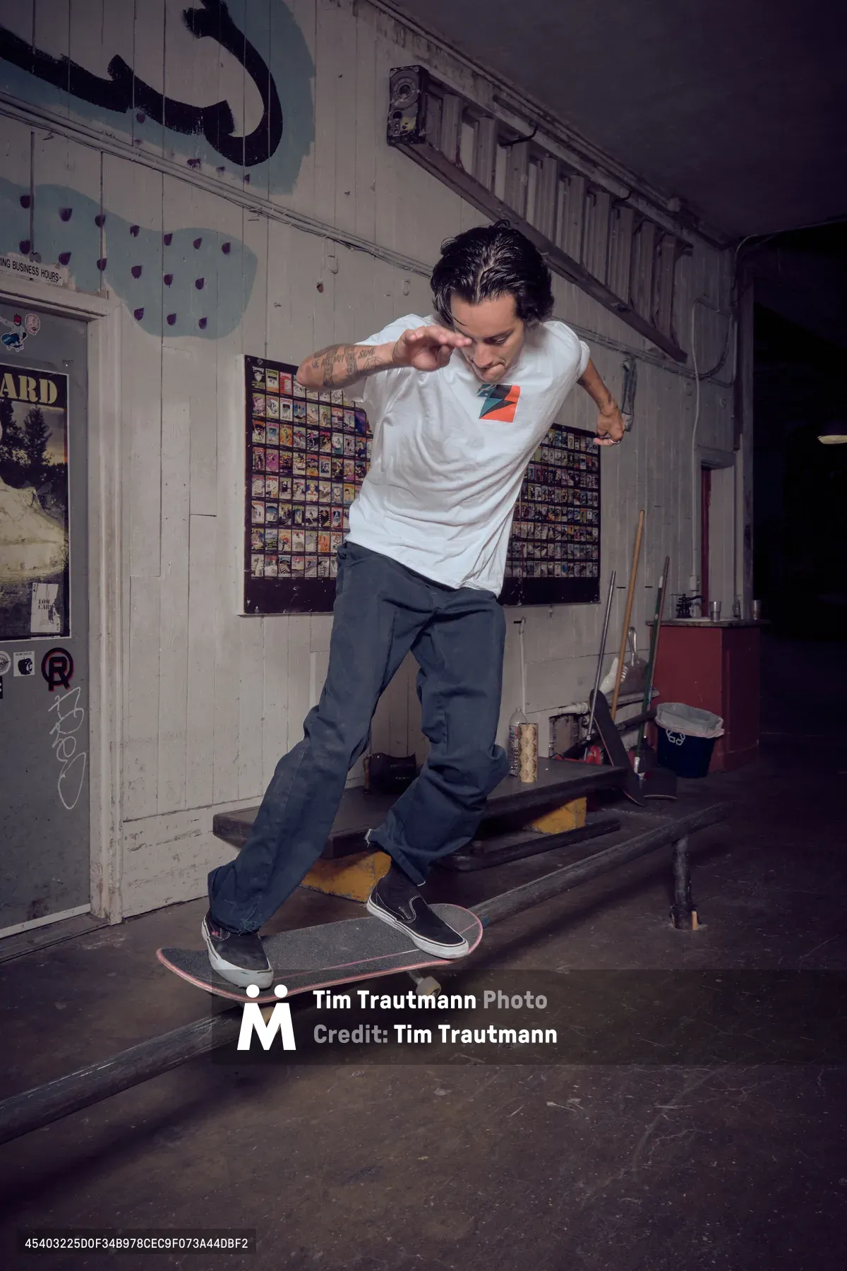 A tattooed skateboarder in a white tee and dark jeans executes a smooth ride through the weathered interior of Commonwealth Skateboarding in Portland's Buckman district. The moody lighting illuminates vintage skate memorabilia plastered across white wooden walls, while the rider's focused expression and fluid motion capture the authentic spirit of skate shop culture. Shadows dance across the worn floorboards as the session unfolds in this iconic Southeast Portland skate sanctuary.
