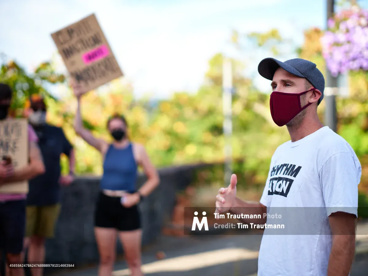 Organizer Peter Marks, wearing a burgundy face mask and gray baseball cap, gestures emphatically while speaking to assembled protesters outside Representative Kurt Schrader's office in Oregon City. The afternoon light casts warm tones across the scene as blurred demonstrators hold signs in the background, creating an atmosphere of peaceful civic engagement. His white t-shirt bearing partial text suggests grassroots activism, while the suburban setting frames this moment of political discourse.