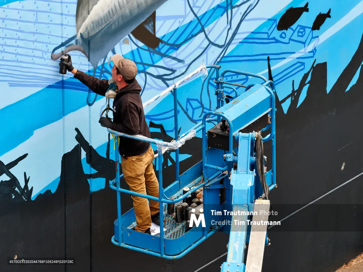 Jeremy Nichols and Devin Finley work from a blue scissor lift to paint an expansive mural featuring flowing blue forms and black silhouettes on a building wall in Southeast Portland's Central Eastside. The artist in mustard-colored pants extends his spray can toward the dynamic composition while bottles of paint rest in the lift's platform. Crisp daylight illuminates the industrial setting where street art transforms urban architecture.