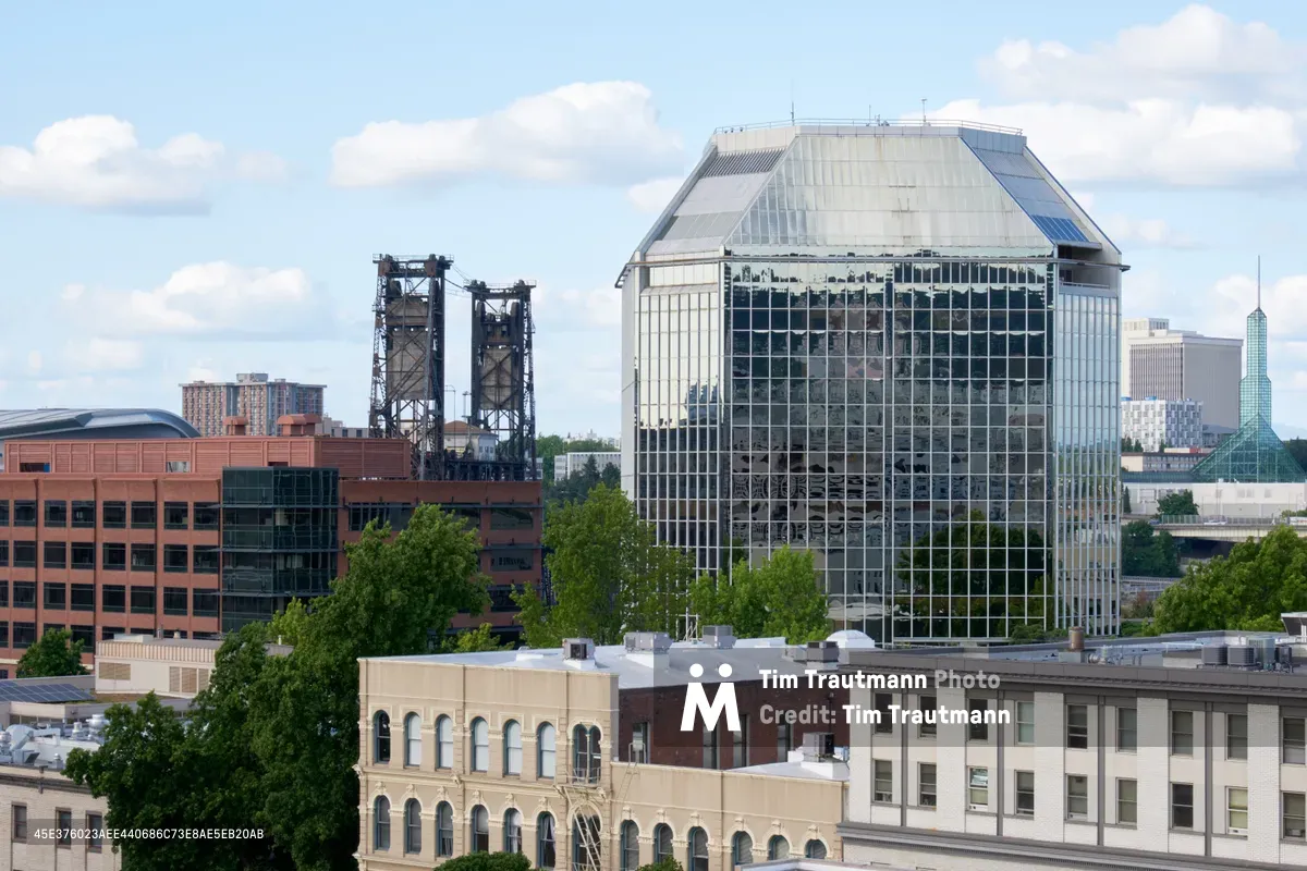 A striking glass conservatory with Victorian-inspired architecture dominates the Portland skyline, its crystalline greenhouse structure gleaming against scattered white clouds. The image captures the juxtaposition of this horticultural landmark against the industrial backdrop of the Fremont Bridge's steel towers and the modern downtown core. Historic brick buildings anchor the foreground, their weathered facades telling stories of Old Town's commercial past, while lush summer foliage softens the urban edges.