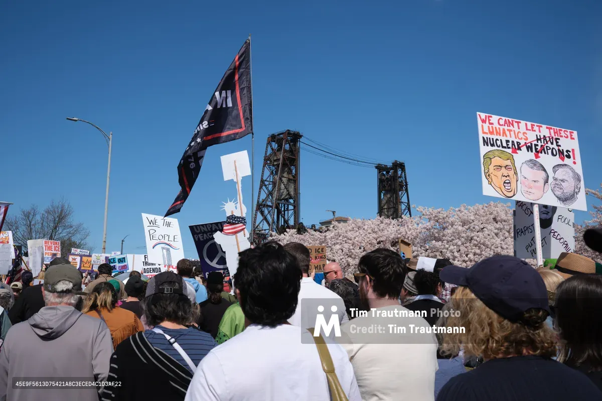 Beneath the iconic steel lattice of Portland's Burnside Bridge, hundreds of demonstrators surge through Northwest Naito Parkway during the March 2026 No Kings Protest. Political signs pierce the crystalline blue sky, including prominent displays warning against nuclear weapons and featuring caricatures of political figures. The crowd flows like a human river between the industrial waterfront and blooming cherry trees, their varied signs and banners creating a tapestry of dissent against the backdrop of Portland's historic Old Town district.