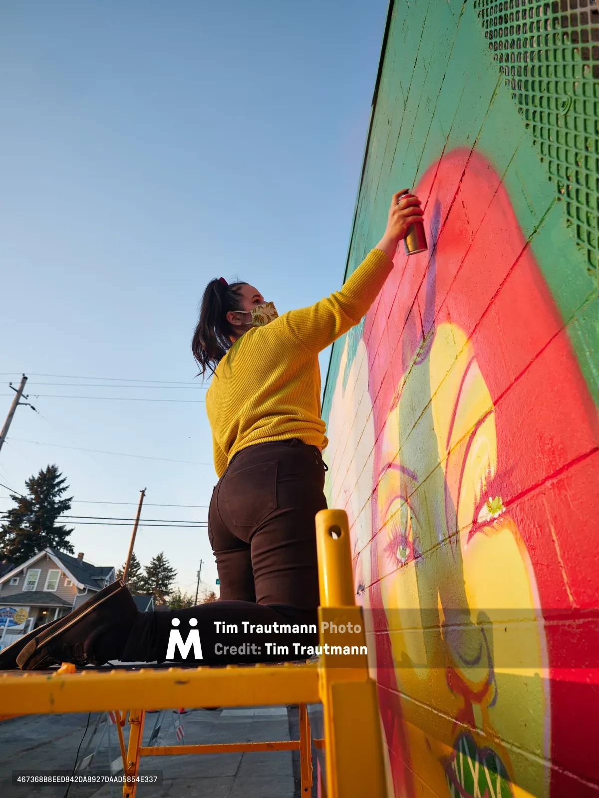 Multidisciplinary artist Villatose stretches upward from a yellow scaffold, spray can in hand, adding coral and yellow brushstrokes to her mural 'Jardín Cubana' depicting three female figures on Northeast Alberta Street in Portland's historic arts district. The golden hour light bathes her mustard sweater and the building's mint green facade, while residential rooflines and power lines frame the creative transformation of this urban canvas. Her focused gesture captures the intersection of community art and neighborhood revitalization in one of Portland's most culturally significant corridors.