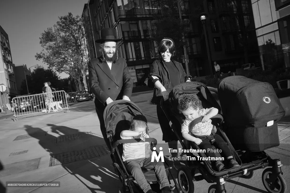 A Hasidic couple guides their double stroller through the sun-dappled sidewalks of Williamsburg, Brooklyn, their traditional black attire creating stark silhouettes against the urban landscape. The father, wearing his characteristic black hat and coat, beams with quiet joy while the mother, in modest dress and sunglasses, tends to their young children nestled in the modern stroller. Dramatic shadows stretch across the concrete as pre-war tenements with fire escapes tower overhead, capturing the intersection of timeless tradition and contemporary city life along Kent Avenue.