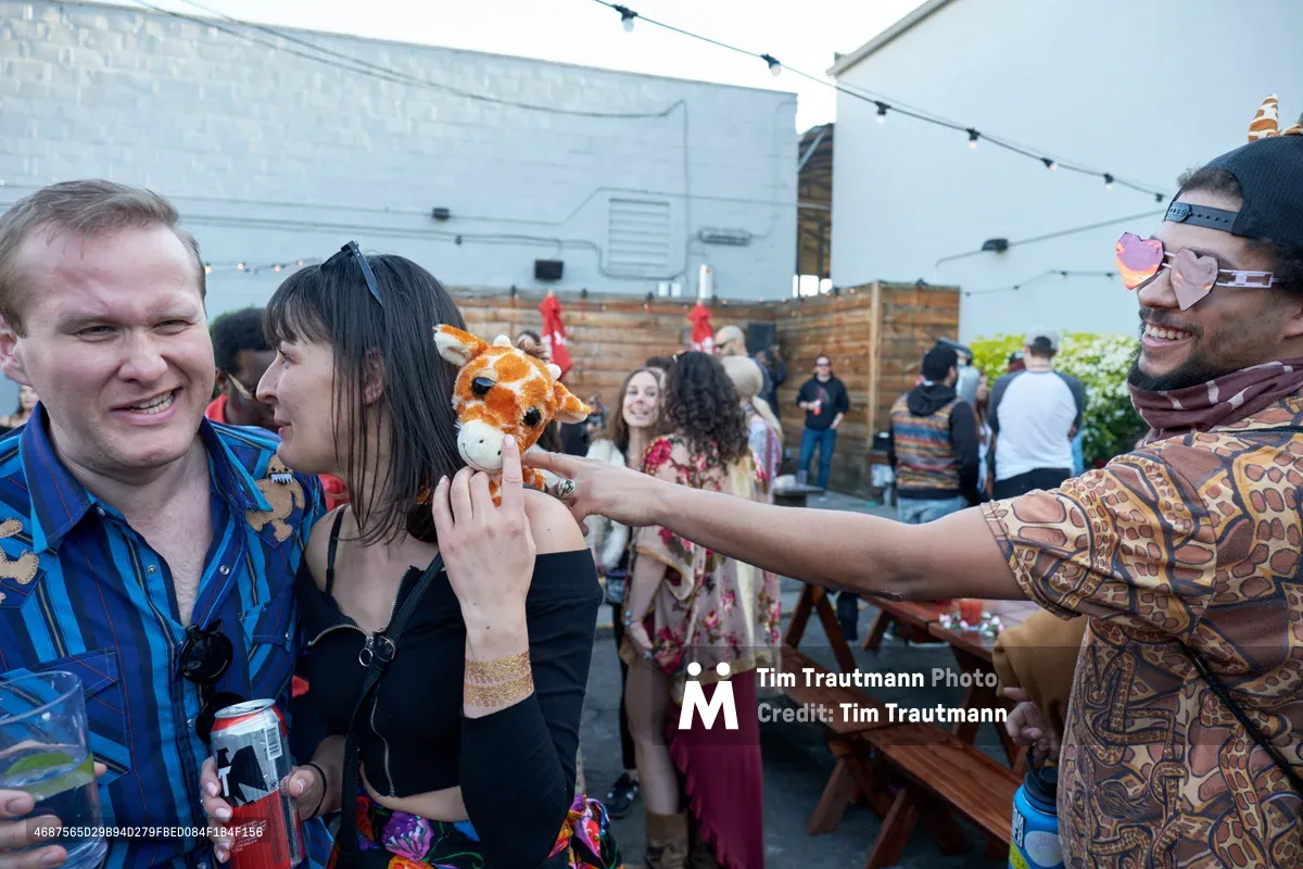 Under the dappled shade of a white canopy at White Owl Social Club, a spirited moment unfolds as a man in heart-shaped sunglasses and paisley shirt playfully extends a plush giraffe puppet toward a woman in black, while her companion in a tropical blue shirt grins with delight. The rooftop gathering buzzes with Portland's characteristic summer energy, string lights and wooden fencing creating an intimate urban oasis where whimsy meets community. The spontaneous puppet theater captures the essence of Your Sunday Best's season opener—unexpected joy blooming in the afternoon light.