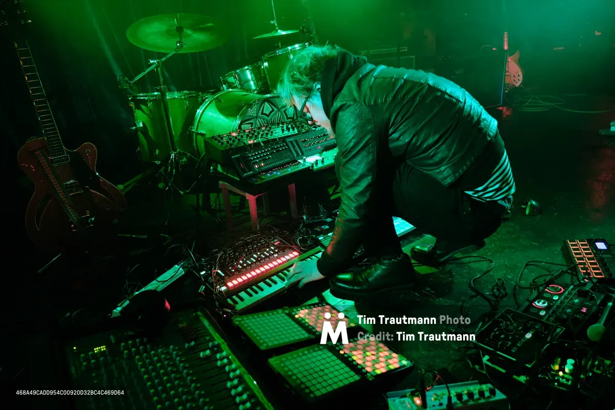 Electronic musician Eli Savage, performing as ellipsism, hunches intently over a sprawling array of synthesizers, drum machines, and MIDI controllers during his live set at RXFest 2019. Bathed in otherworldly emerald stage lighting, the artist manipulates his electronic instruments with focused precision while surrounded by the intimate darkness of Dante's venue in Portland's downtown core. The atmospheric green wash transforms the performance space into a cyberpunk dreamscape, highlighting the glowing control surfaces and creating dramatic shadows across the cluttered stage floor.