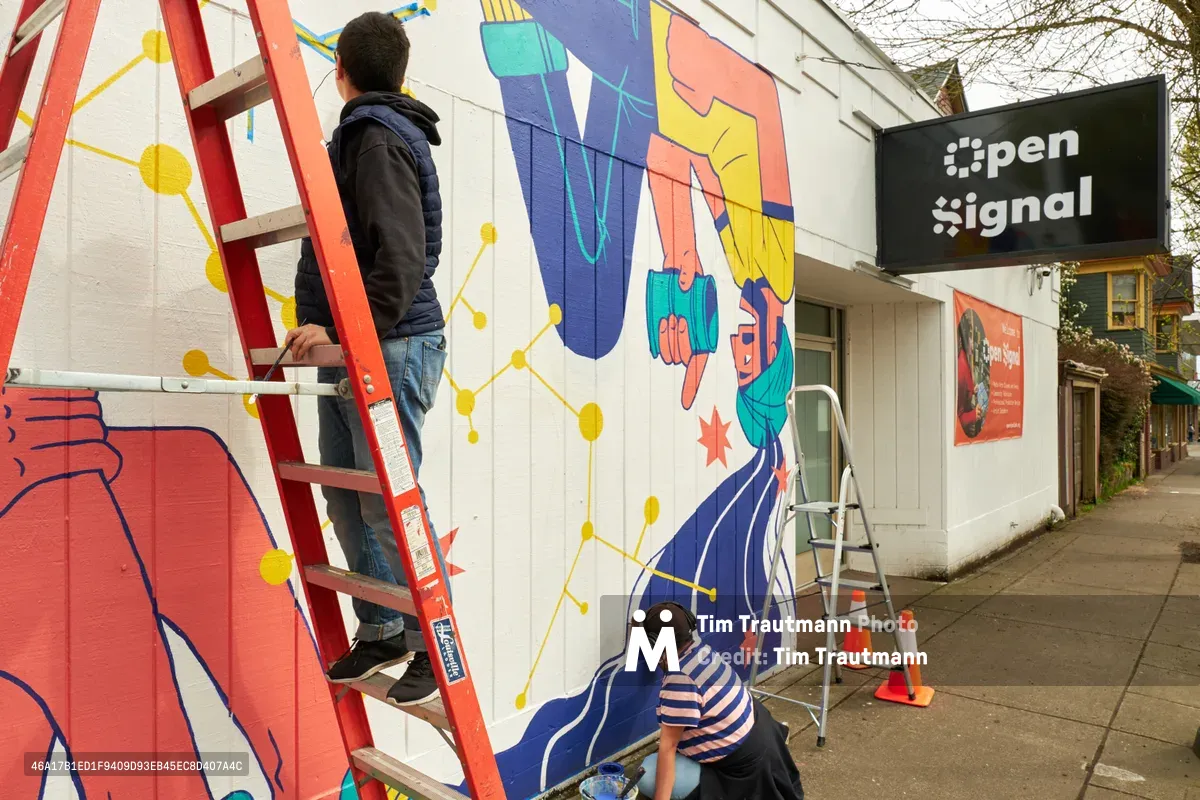Two muralists work collaboratively on a vibrant street art installation outside Open Signal community media center in Portland's Eliot neighborhood. One artist perches on a red ladder while another crouches at ground level, both adding colorful details to the large-scale public artwork featuring abstract figures and geometric patterns in blues, yellows, and coral tones. The scene captures the intimate process of community art creation, with painting supplies scattered on the sidewalk and the artists' focused concentration visible as they bring their vision to life on the white brick wall.