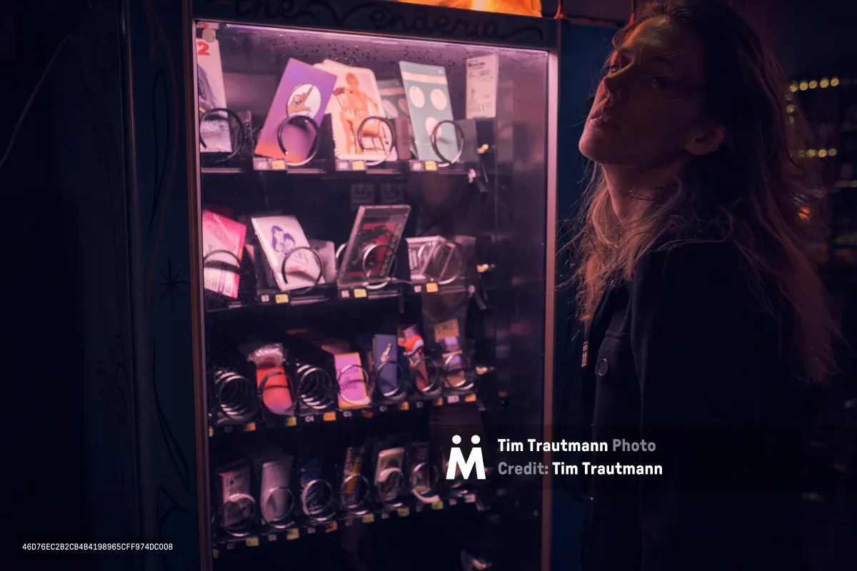 A young woman with long dark hair gazes upward at an illuminated vending machine in The Florida Room on North Killingsworth Street in Portland's Humboldt neighborhood. The machine's internal pink and purple fluorescent lighting creates a nostalgic glow against the surrounding darkness, displaying rows of packaged goods behind glass. Her profile is softly lit by the machine's ambient light, capturing a moment of late-night contemplation and urban solitude.
