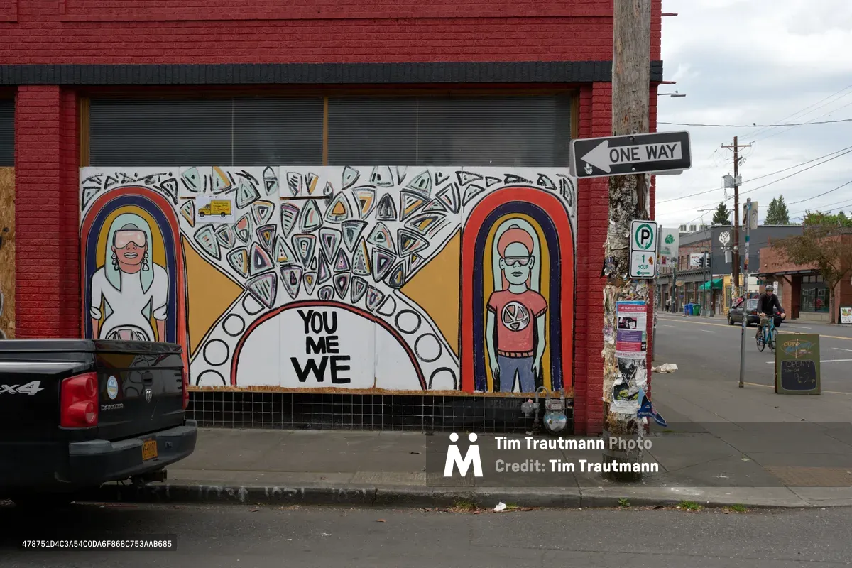 A vibrant mural depicting two figures within rainbow arcs and the message 'YOU ME WE' adorns the storefront of a shuttered business on Southeast Hawthorne Boulevard in Portland's Richmond neighborhood. The artwork transforms the reality of economic hardship into a beacon of community solidarity, with intricate geometric patterns radiating from the central message. Overcast skies and quiet streets frame this testament to artistic activism in one of Portland's historically eclectic districts.