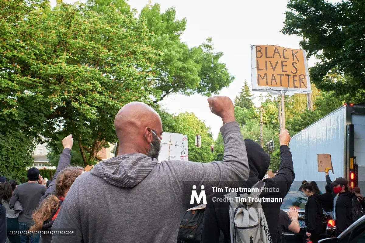 A bald man in a gray hoodie raises his fist skyward while holding a handwritten protest sign during a Black Lives Matter demonstration in Portland's Laurelhurst neighborhood. The crowd fills the tree-lined intersection of Northeast César Chávez Boulevard and East Burnside, their cardboard signs creating a forest of demands for justice beneath an overcast spring sky. Behind the protesters, a blue bus waits in traffic as the peaceful demonstration temporarily claims the urban streetscape.