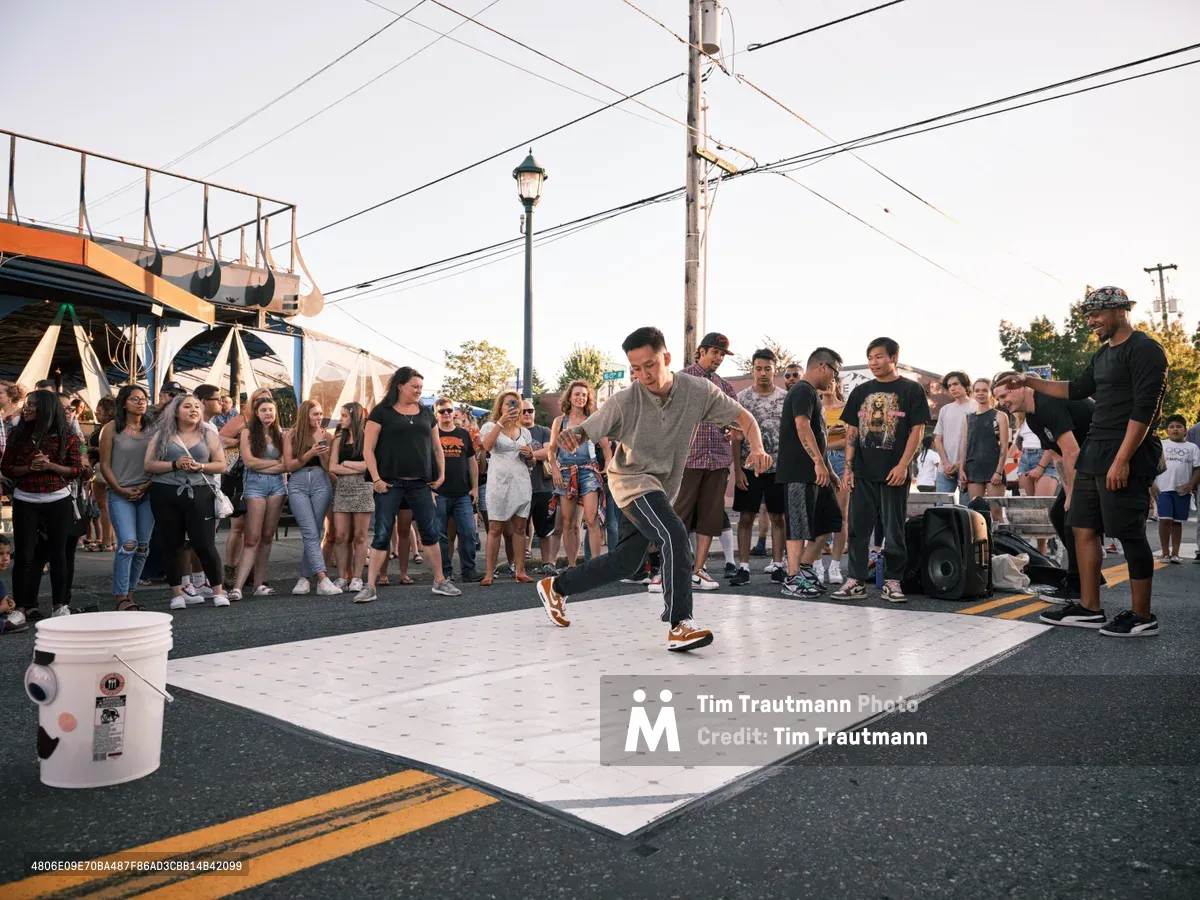 A breakdancer in a grey sweatshirt commands the center of a white dance mat laid out on Alberta Street's asphalt, his body frozen in mid-move as dozens of spectators form a tight semicircle around the performance space. The golden hour light bathes the scene in warm tones while power lines crisscross overhead, creating an urban canopy above this spontaneous street performance at Portland's Last Thursday event. Young faces lean forward with anticipation, some recording on phones, as the energy of Northeast Portland's vibrant arts district pulses through the gathered crowd.