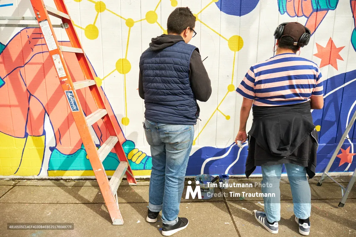 Two muralists work on the vibrant "Let's Talk" public art installation at Open Signal in Portland's Eliot neighborhood. One artist in a navy vest and hoodie stands contemplatively before the wall while another in a striped shirt and headphones actively paints the colorful geometric design. The mural features bold yellow molecular patterns, coral and teal geometric shapes, and dynamic blue elements against a white concrete wall, with an orange stepladder and paint supplies scattered across the concrete sidewalk.