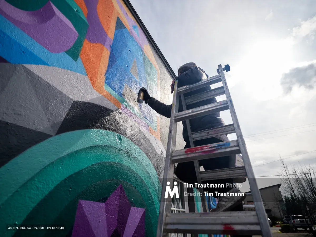 A street artist perched on an aluminum ladder applies bold strokes to a kaleidoscopic mural bursting with turquoise, purple, and orange geometric forms. Shot from a low angle against Portland's overcast sky, the composition emphasizes the scale and ambition of this urban art project in the northeast district. The artist's focused concentration and the mural's flowing, abstract patterns create a dynamic tension between human creativity and architectural canvas.