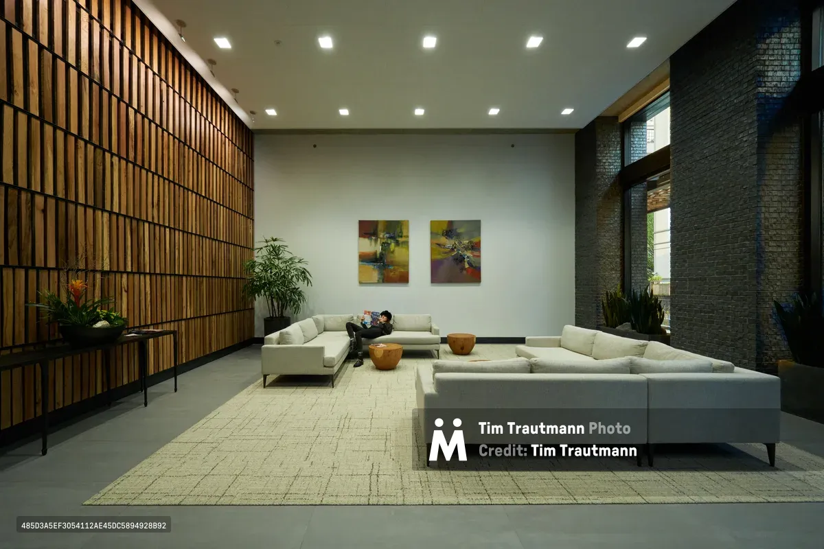A solitary figure reclines on pristine white modular seating in the sophisticated lobby of Pearl West, a contemporary residential building in Portland's Pearl District. The space showcases striking architectural contrasts between warm vertical wood slats and cool charcoal brick, while recessed ceiling lights cast an even glow across polished concrete floors. Abstract artwork punctuates the neutral palette as the resident finds quiet respite in this carefully curated urban sanctuary.