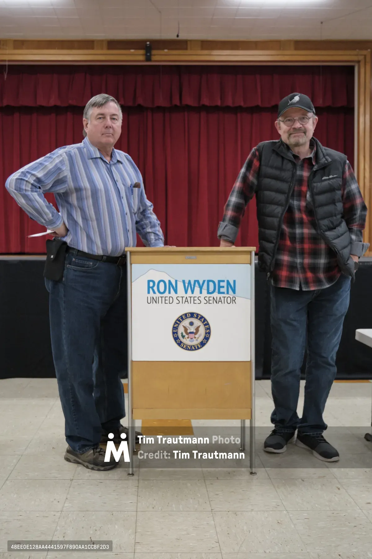 Two middle-aged photographers representing the American Society of Media Photographers stand casually beside a Ron Wyden United States Senator podium in what appears to be a school gymnasium or community center. The man on the left wears a blue striped dress shirt with jeans, while his colleague on the right sports a plaid flannel shirt under a dark vest with a baseball cap. Behind them, burgundy curtains frame the scene against institutional white tile ceiling and polished concrete floors, creating an atmosphere of civic engagement in Portland's Robert Gray Middle School.