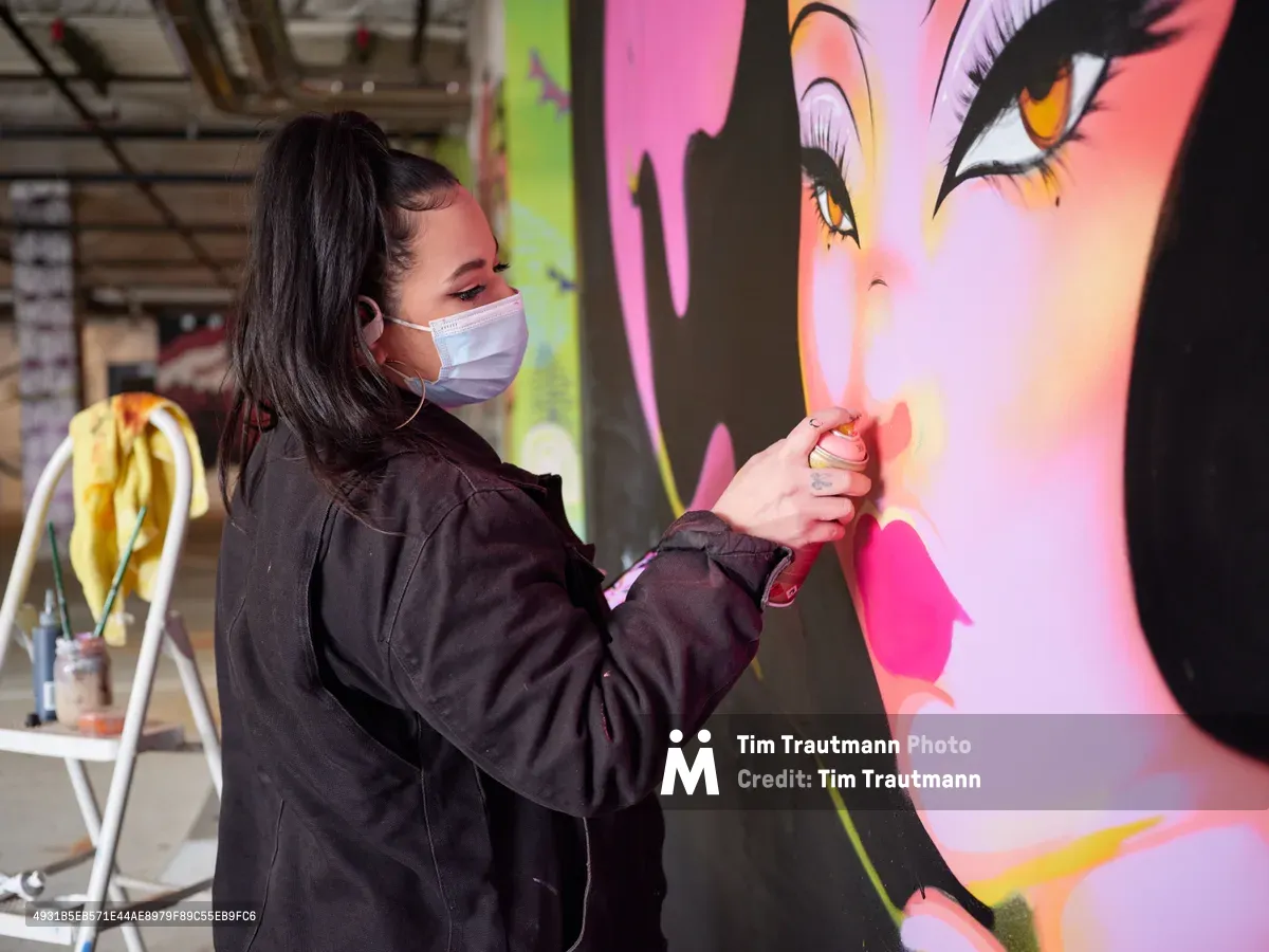 Multidisciplinary artist Vilatose applies finishing touches to a luminous portrait mural in the industrial confines of Clay Creative's parking garage in Portland's Hosford-Abernethy neighborhood. Wearing a protective mask and dark work attire, she delicately airbrush details onto the large-scale feminine face rendered in electric pinks, corals, and sunset hues. The contrast between the raw concrete surroundings and the radiant artwork creates a compelling dialogue between urban grit and artistic transformation.