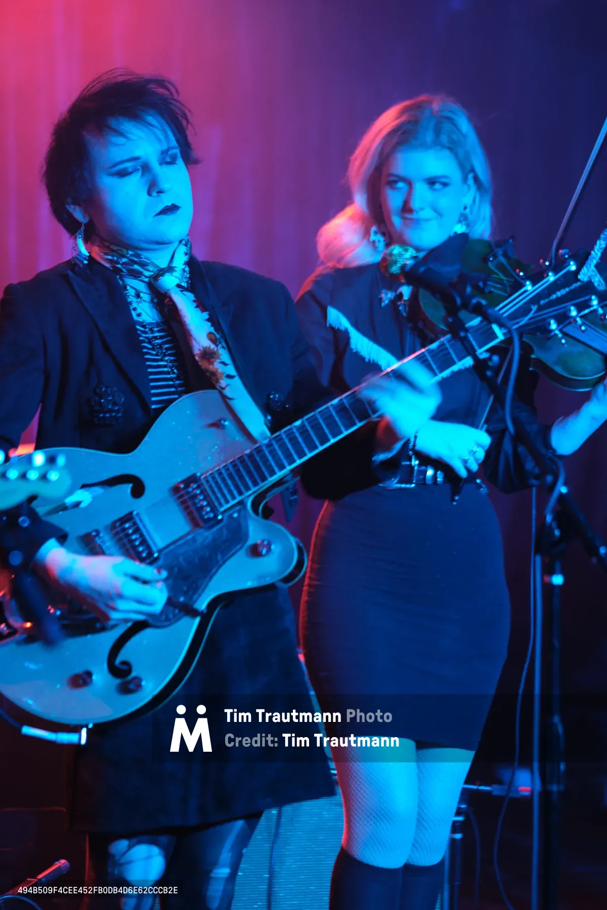Charlotte McCaslin and Faith Grossnicklaus of Roselit Bone command the stage at Dante's in Portland, bathed in electric blue and magenta light. McCaslin's white hollow-body guitar gleams under the theatrical lighting while Grossnicklaus draws her violin bow with focused intensity. The atmospheric haze and saturated color wash transforms the intimate venue into a dreamlike sonic landscape, capturing the duo's genre-blending artistry in full flight.