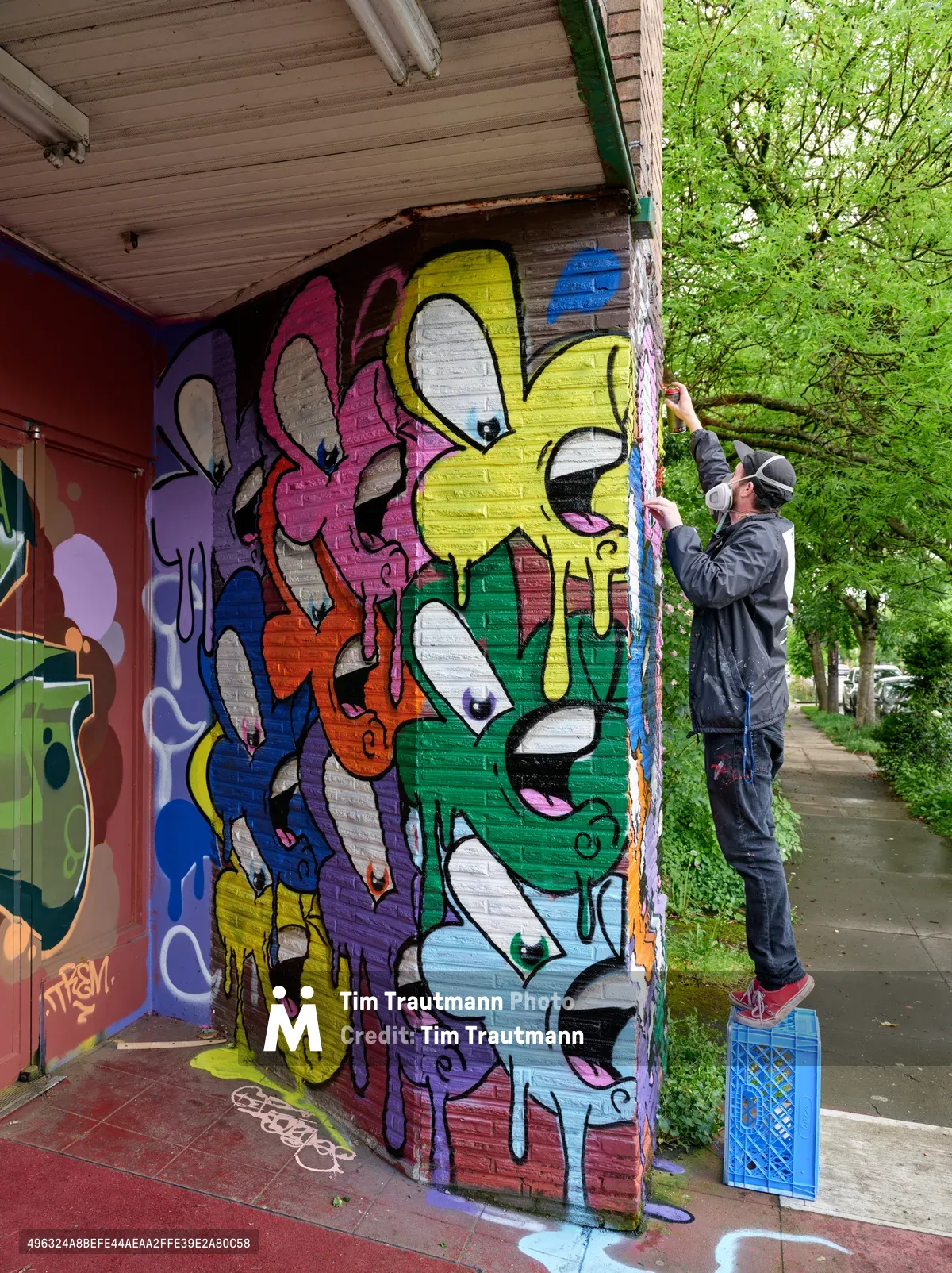 A street artist stands on a blue milk crate, meticulously adding details to a kaleidoscopic mural adorning the brick facade of the former Oregon Theater on Southeast Division Street in Portland's Richmond neighborhood. The composition bursts with cartoon-like characters in electric yellows, magentas, blues, and greens, their bulbous forms creating a playful contrast against the weathered brick and wooden eaves. Lush spring foliage frames the scene, while the artist's focused concentration captures the intimate process of transforming urban architecture into community art.
