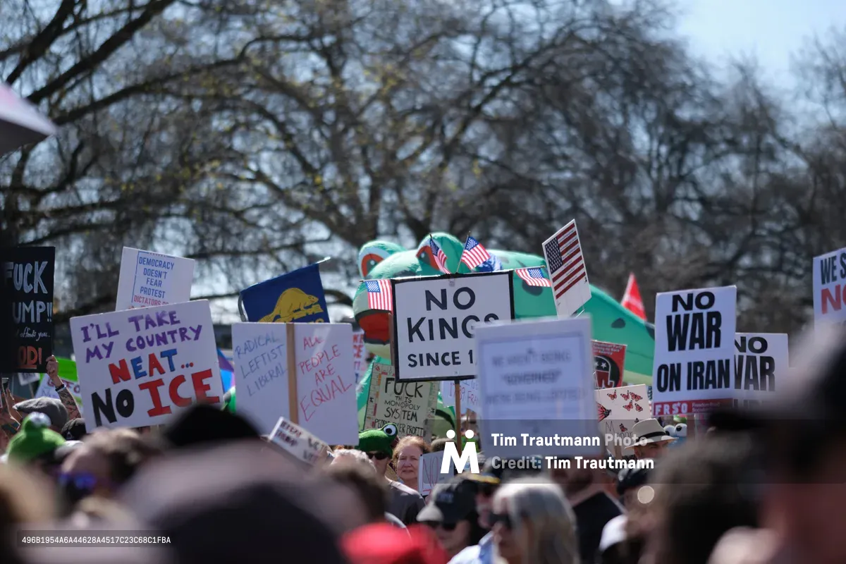 A sea of protest signs rises above a dense crowd gathered in Portland's historic Old Town district, with bare winter trees creating a skeletal canopy overhead. Hand-lettered placards declare "NO KINGS SINCE 1" and "NO WAR ON IRAN" while American flags punctuate the political messaging, creating a tapestry of democratic dissent. The shallow depth of field blurs the crowd into an anonymous mass while sharp focus on the signs emphasizes their urgent messages. Overcast Pacific Northwest skies cast even, diffused light across the demonstration, lending gravity to this moment of civic engagement.
