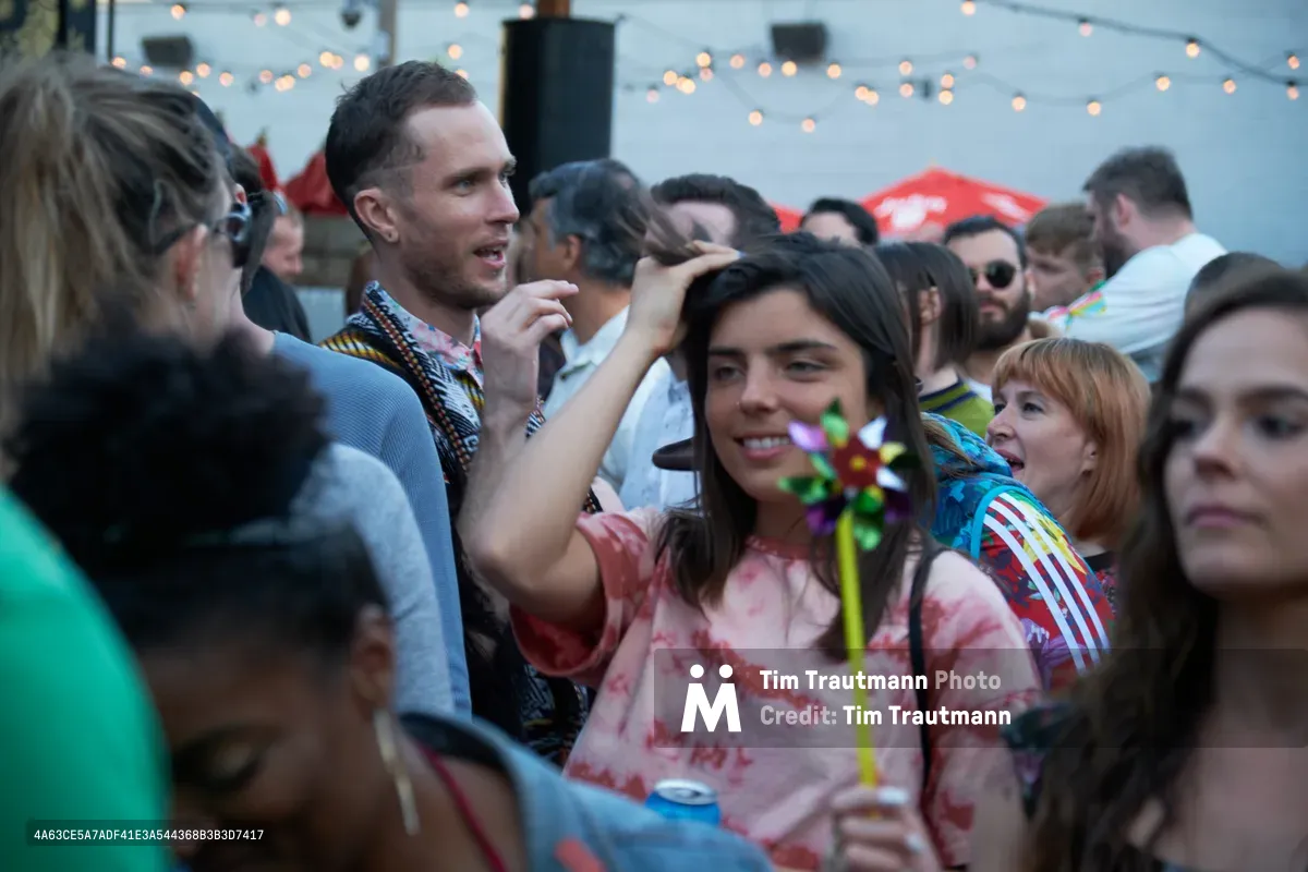 A jubilant crowd gathers at White Owl Social Club during Your Sunday Best's season opener, their faces illuminated by warm string lights overhead. Festival-goers sport vibrant tie-dyed shirts and casual summer attire, creating an intimate tapestry of celebration in Portland's beloved outdoor venue. The shallow depth of field captures genuine moments of connection as friends laugh and socialize in the golden hour glow.