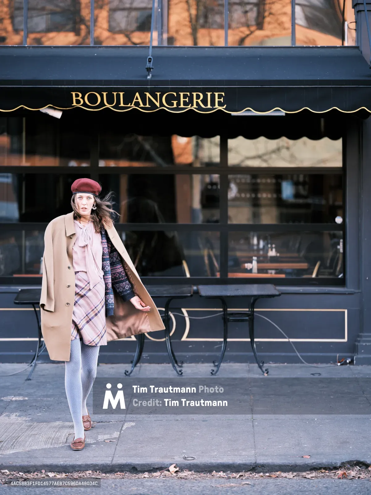 A young woman in a burgundy beret and camel coat strikes a dynamic pose outside a boulangerie on Portland's Alberta Street. Her wind-swept hair and layered ensemble of plaid scarf, patterned sweater, and white tights create an effortlessly Parisian aesthetic against the urban backdrop. The golden afternoon light catches the brass lettering of the French bakery behind her, while outdoor café chairs hint at European-style street life in this Northeast Portland neighborhood.