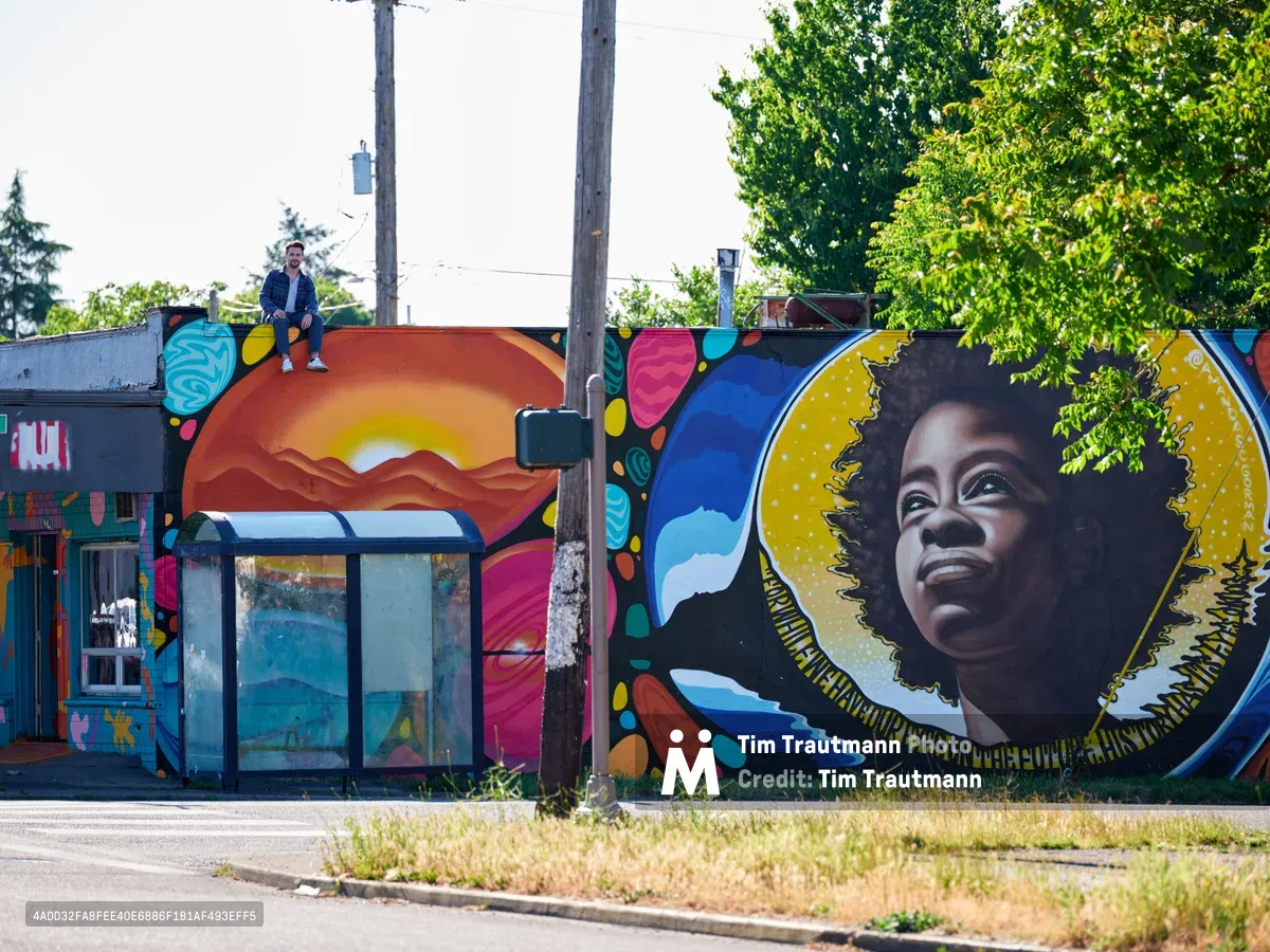 Travis Fields sits triumphantly atop a weathered building in Portland's Alberta district, having completed a vibrant mural celebrating poet Amanda Gorman. The large-scale artwork features Gorman's portrait rendered with warm golden tones against a backdrop of sunset mountains and swirling celestial elements. Fields, positioned on the building's edge beneath overcast skies, embodies the satisfaction of artistic completion while power lines and verdant summer foliage frame this moment of creative achievement in Northeast Portland.