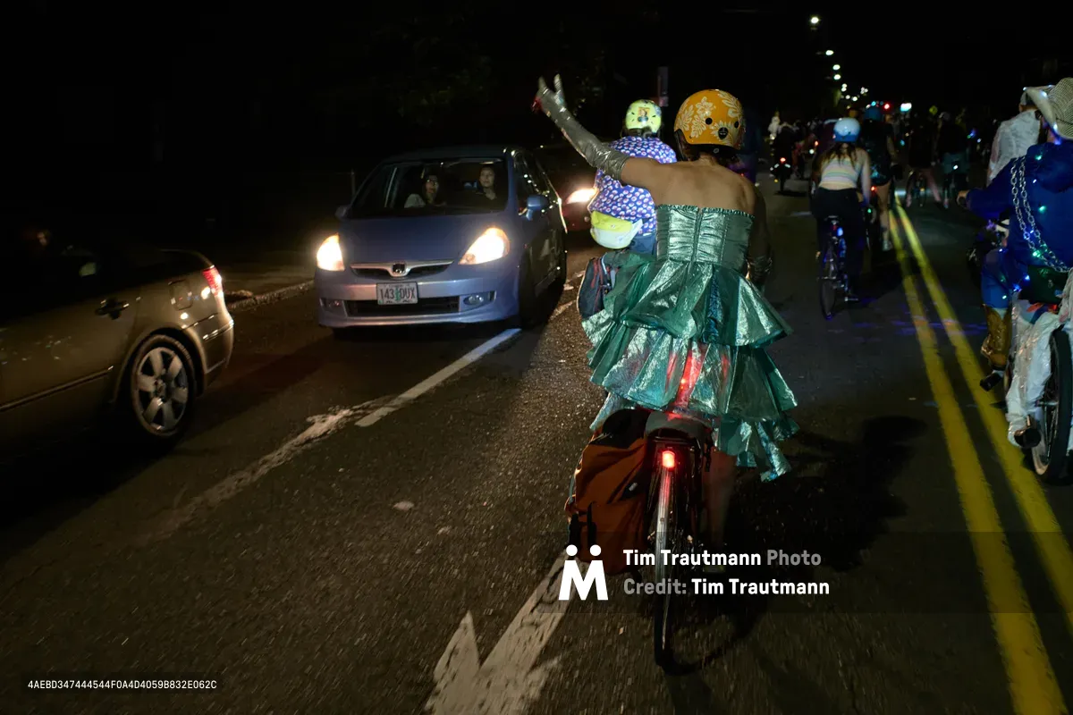 A whimsical nighttime procession unfolds along Southeast Lincoln Street as costumed cyclists transform Portland's asphalt into a makeshift ballroom. Two riders in elaborate formal wear—one sporting a shimmering teal gown, the other in polka-dotted attire—navigate between curious motorists with theatrical flair. The scene captures the anarchic spirit of Pedalpalooza's Drop Out Prom Ride, where bicycle culture collides with formal rebellion under the glow of headlights and street lamps.