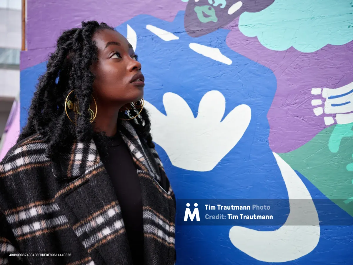 Artist Naomi Likayi stands in contemplative profile against her vibrant mural at Portland's boarded World Trade Center, her natural locs framing her upward gaze. The kaleidoscopic backdrop features flowing abstract forms in electric blues, purples, and mint greens with playful white hand motifs. Golden hoop earrings catch the light as she wears a geometric plaid coat, creating an intimate portrait that bridges the artist with her transformative public art commissioned by the Portland Street Art Alliance.