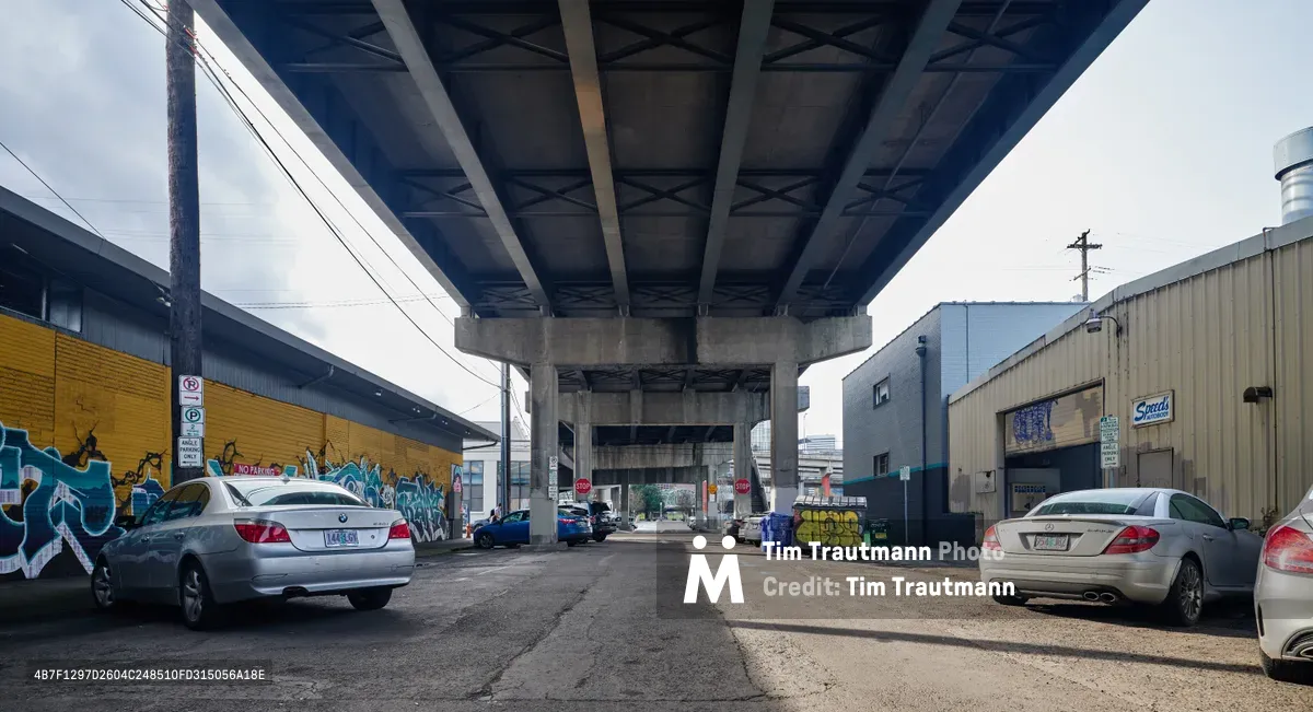 A weathered street scene unfolds beneath Portland's concrete overpass in the Central Eastside district, where commissioned street murals bloom against warehouse walls. Silver sedans and a luxury coupe rest on cracked asphalt, framed by the industrial geometry of bridge supports and power lines. The muted daylight filters through overcast skies, casting the urban corridor in soft, diffused tones that emphasize the textural contrast between raw concrete infrastructure and vibrant graffiti art.
