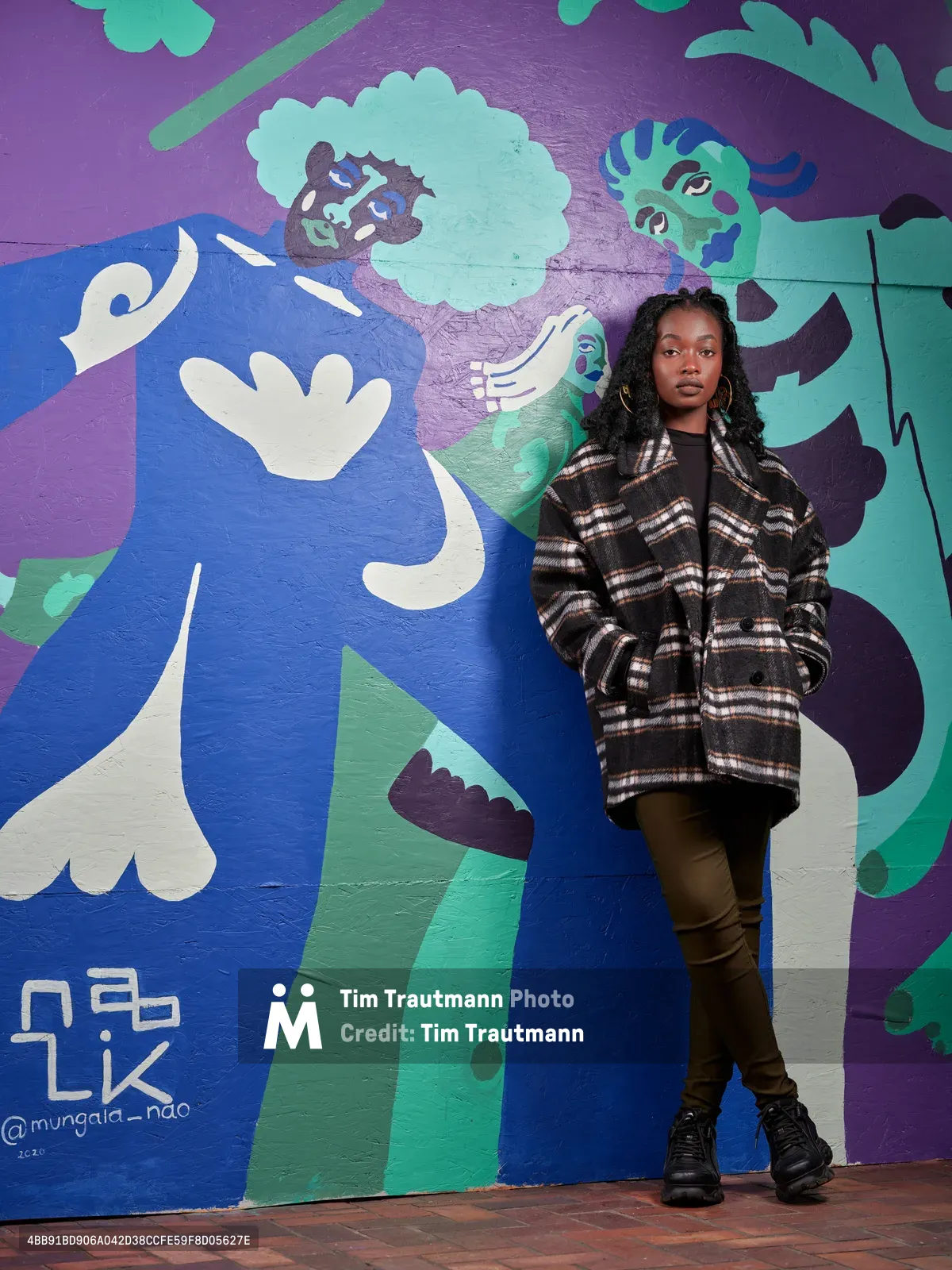 Naomi Likayi stands confidently against her vibrant mural painted on the boarded Portland World Trade Center, commissioned by the Portland Street Art Alliance. The artist wears a plaid coat and poses with hands in pockets beside her abstract work featuring blues, purples, and greens with stylized figures and organic shapes. The urban street art transforms the temporary boarding into a canvas of community expression, with the artist's signature visible in the corner of her colorful composition.
