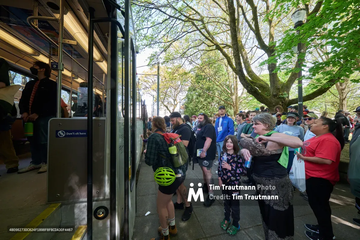 A diverse crowd of transit enthusiasts and families gathers around TriMet's retiring Type 1 MAX light rail car during its farewell event at Holladay Park in Portland's Lloyd District. The spring afternoon light filters through mature trees as passengers board for one last ride on the historic vehicle that has served the city since the system's inception. Children and adults alike press close to the train, their faces reflecting both nostalgia and curiosity as they witness the end of an era in Portland's public transportation history.