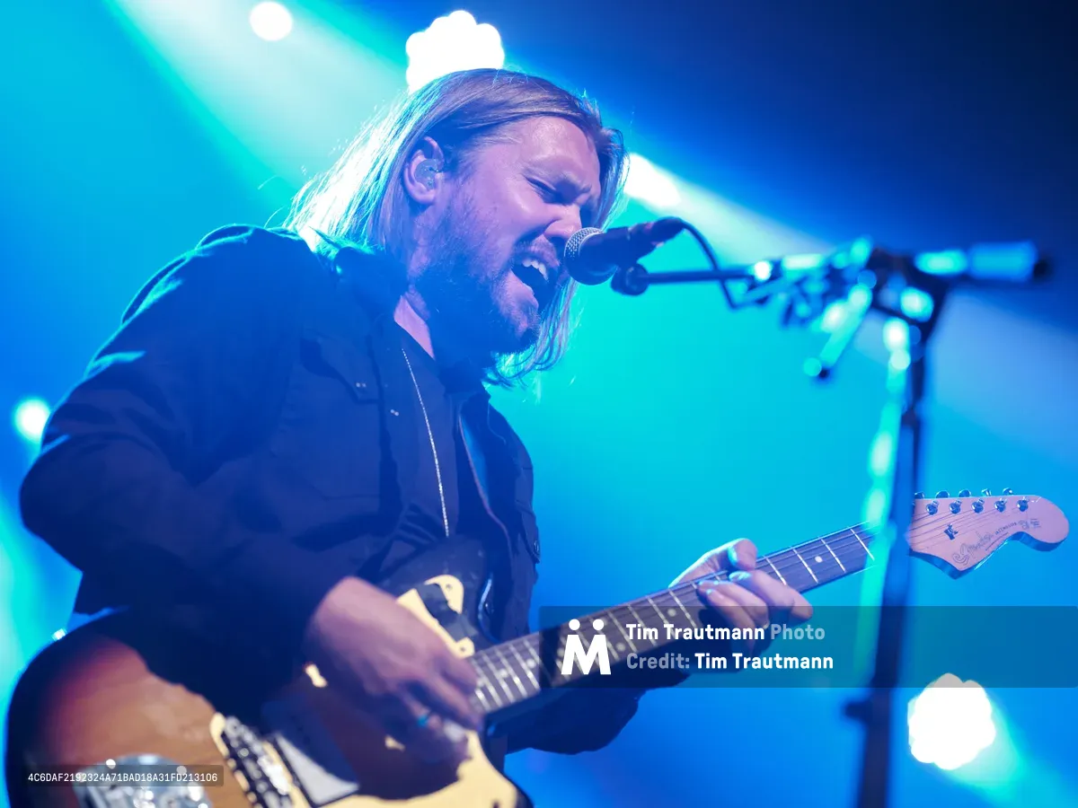 Russell Marsden of Band of Skulls delivers an intense vocal performance while gripping his electric guitar during the band's 10 Year Anniversary Tour at Webster Hall in Manhattan. Dramatic stage lighting bathes the long-haired musician in ethereal blue and cyan hues, creating a dreamlike atmosphere as he leans into the microphone with passionate intensity. The iconic venue's professional lighting rig creates brilliant bokeh effects in the background, while Marsden's focused expression and dynamic pose capture the raw energy of live rock performance.