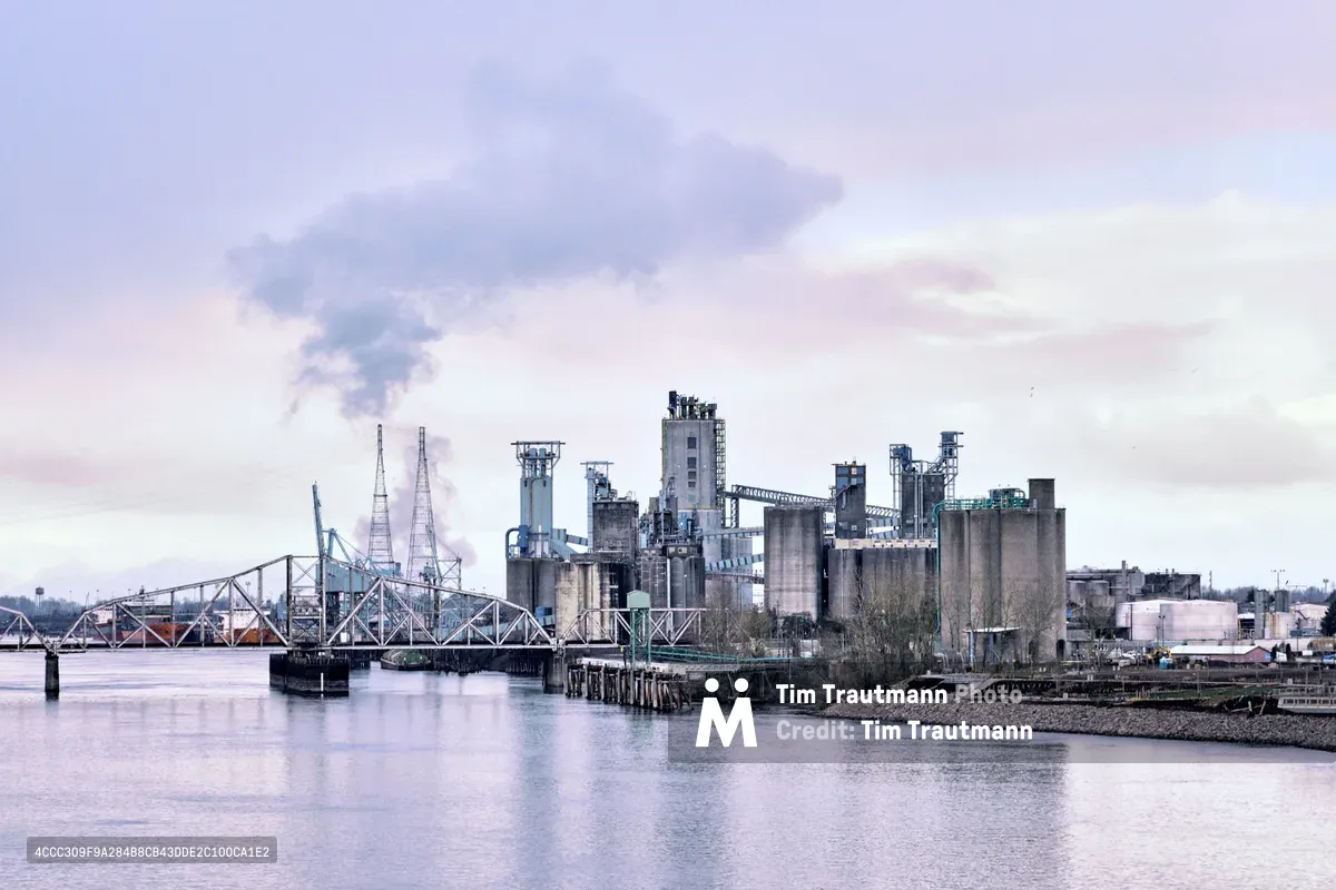 The Great Western Malting facility rises like a concrete fortress along the Washington shoreline of the Columbia River, its grain silos and processing towers silhouetted against a pearl-gray afternoon sky. Steam wisps from the industrial complex while a steel truss bridge spans the glassy water, creating perfect reflections in the still surface. The scene captures the intersection of heavy industry and natural beauty, where one of North America's few ocean-accessible malt plants processes grain destined for global markets.
