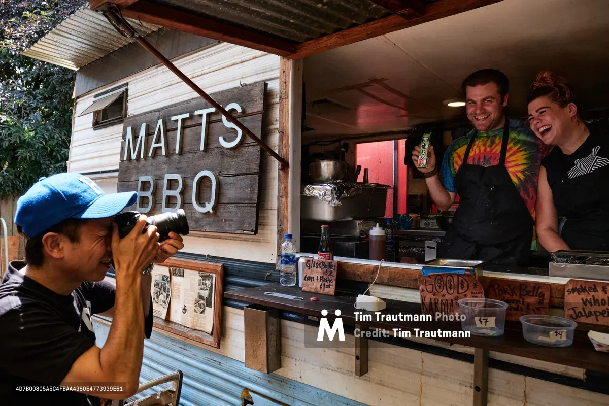 A Japanese tourist wearing a blue baseball cap photographs two laughing staff members at Matt's BBQ food cart on Mississippi Avenue in Portland, Oregon. The male staff member in a tie-dye shirt and black apron holds up a small Japanese snack item for the camera, while his female colleague beside him laughs. The rustic wood-sided cart displays a hand-lettered sign reading "Matt's BBQ," along with menu signs for pork belly and smoked jalapeño. Handwritten signs on the counter read "Good Karma" and "Glass Bottles $2."