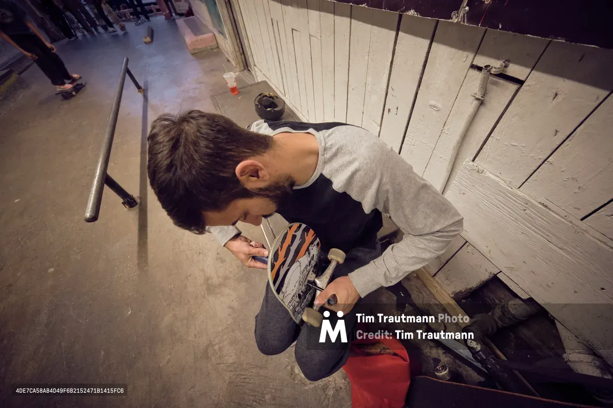 A young skateboarder crouches in concentrated focus, carefully adjusting his board's trucks in the industrial confines of Commonwealth Skateboarding in Portland's Buckman neighborhood. The warm overhead lighting casts dramatic shadows across the weathered wooden ramp structure, while concrete floors and metal railings frame this intimate moment of preparation. His gray hoodie and dark jeans blend with the utilitarian atmosphere of this Southeast Portland skate sanctuary, where craft meets passion.