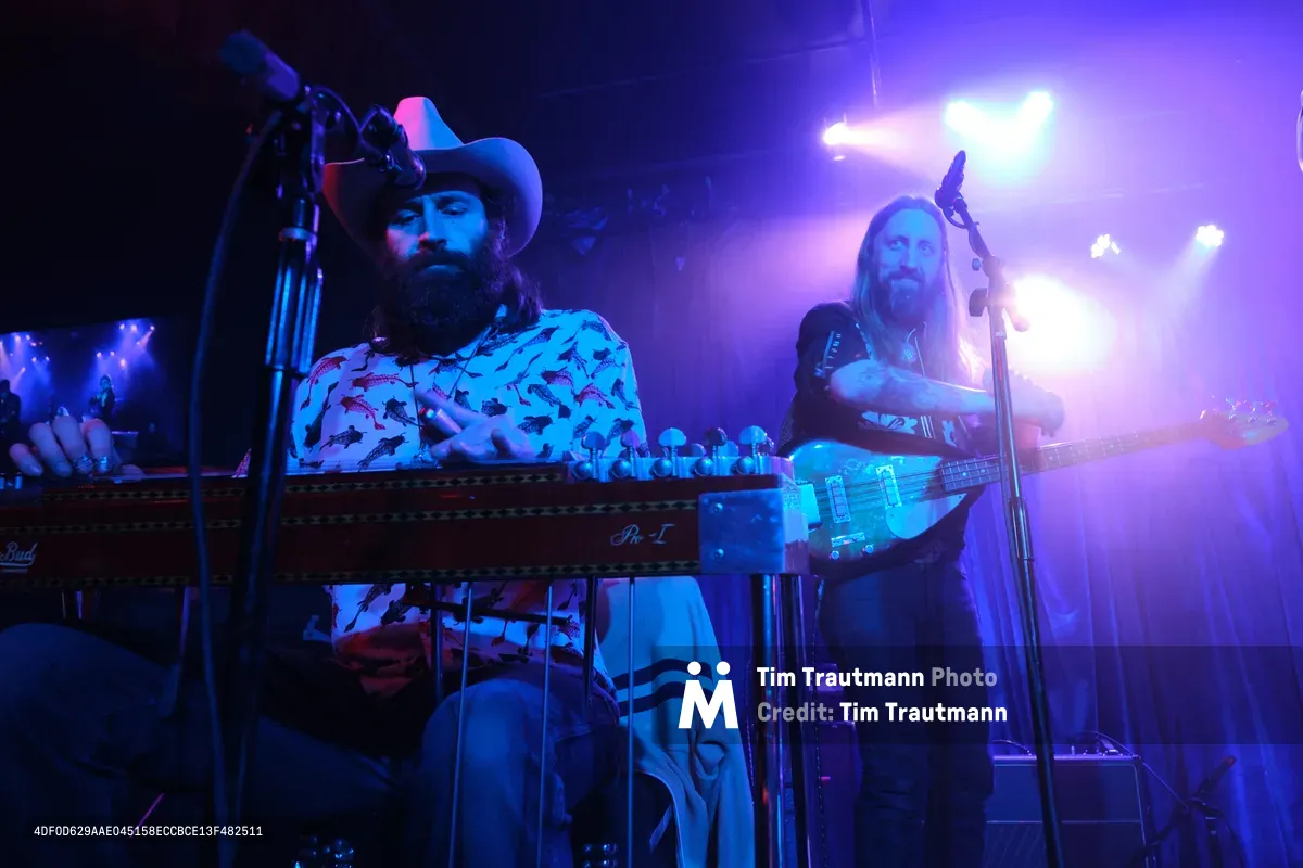 Barry Walker commands the keyboard while Tobias Berblinger anchors the rhythm on bass as Roselit Bone delivers an electrifying performance at Dante's in Portland, Oregon. The duo is bathed in saturated blue and purple stage lighting that cuts through the intimate venue's darkness, creating an atmospheric backdrop for their musical collaboration. Walker's cowboy hat and patterned shirt catch the colored light as he leans into his performance, while Berblinger's long hair flows as he works the bass strings. The dramatic lighting transforms the small club into a sonic cathedral, with beams piercing through the smoky air above the musicians.