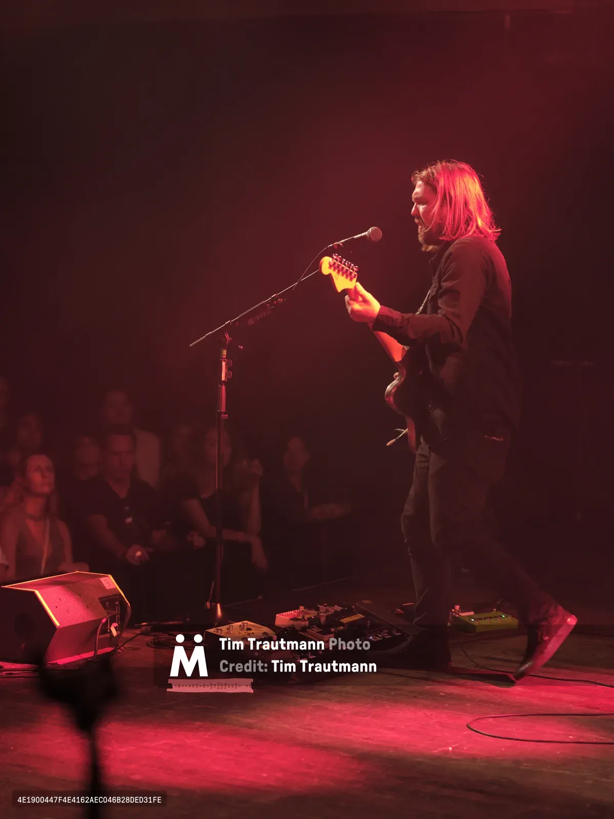 Russell Marsden of Band of Skulls emerges from crimson stage light during the band's 10 Year Anniversary Tour at Manhattan's historic Webster Hall. The guitarist-vocalist leans into his microphone with shoulder-length blonde hair catching the warm red illumination, while an array of effects pedals spreads across the venue's wooden stage floor. The intimate crowd sits in shadows beyond the performance area, creating a striking contrast between the illuminated performer and the darkened audience.