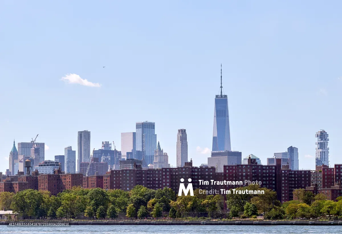 The gleaming spire of One World Trade Center pierces a hazy afternoon sky, dominating Manhattan's Financial District skyline as viewed across the Hudson River. Red-brick housing projects stretch along the waterfront in the foreground, their uniform facades creating a textural contrast against the soaring glass and steel towers beyond. Lush summer foliage softens the urban landscape while a solitary bird glides through the pale blue expanse above the city's architectural symphony.