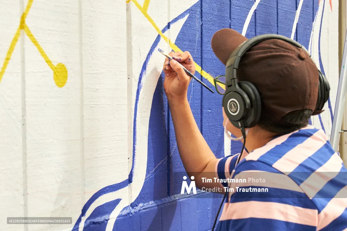 Mexican-American artist Maria Rodriguez, known as Sparkykneecap, works intently on her commissioned mural "Let's Talk" at Open Signal in Portland's Eliot neighborhood. Wearing black headphones and a blue-and-white striped shirt, she applies precise brushstrokes of vibrant blue paint to the white wall surface. The emerging artwork features bold geometric shapes and flowing yellow accents that reflect her signature exploration of identity and cultural themes through playful color and form.