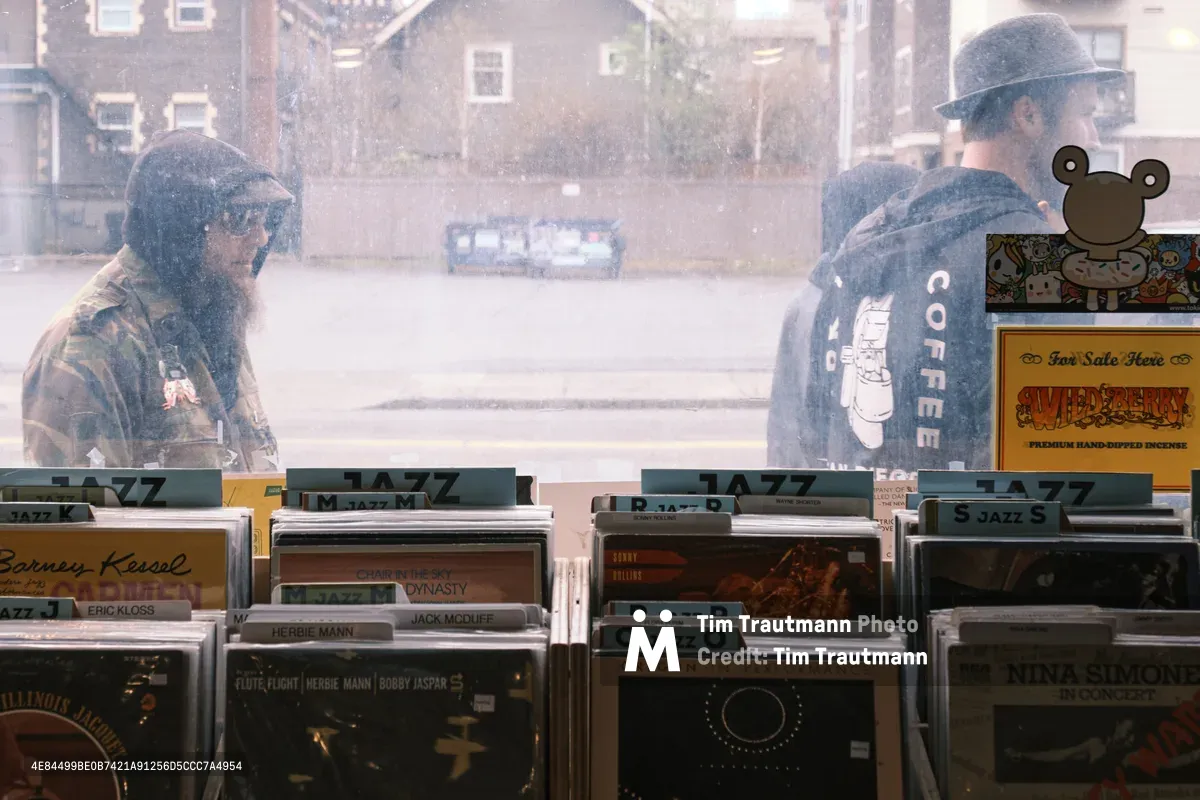 Through the dusty window of a Burnside Street record shop, two hooded figures pass by as vinyl jazz albums sit organized in wooden crates during Record Store Day. The smudged glass creates an intimate, nostalgic quality as records by legends like Nina Simone and Herbie Mann await discovery inside. The weathered window adds character to this quintessential celebration of independently owned record stores.