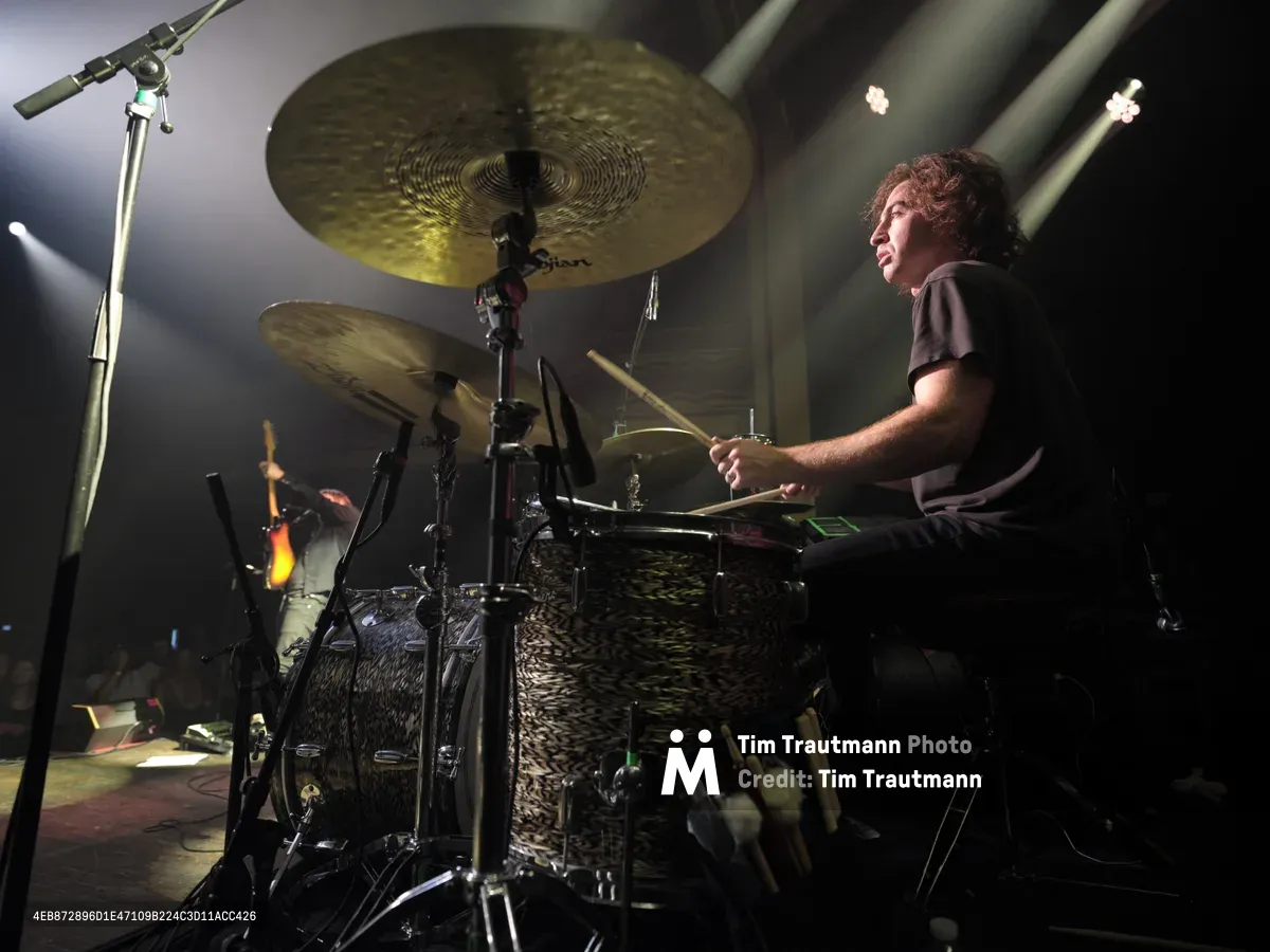 Julian Dorio commands his drum kit during Band of Skulls' 10 Year Anniversary Tour performance at the iconic Webster Hall in Manhattan's East Village. Dramatic stage lighting cuts through the venue's atmospheric haze, illuminating golden cymbals and the drummer's focused expression as he drives the rhythm forward. The intimate concert setting captures the raw energy of live rock performance, with microphone stands and equipment creating layered silhouettes against the moody backdrop.