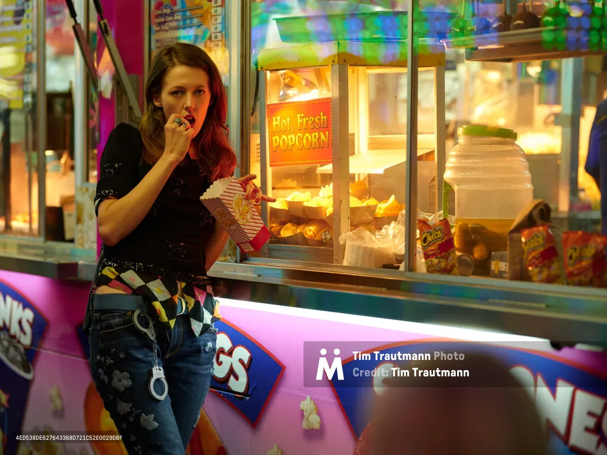 A young woman with auburn hair enjoys popcorn from a striped paper container beside a glowing concession stand at the Clark County Fair in Vancouver, Washington. The warm amber light from the "Hot, Fresh Popcorn" machine creates an inviting atmosphere against the kaleidoscope of carnival colors and neon signage. Her casual attire of distressed jeans and dark top contrasts with the vibrant fair environment, capturing a moment of simple pleasure amid the sensory overload of the midway.