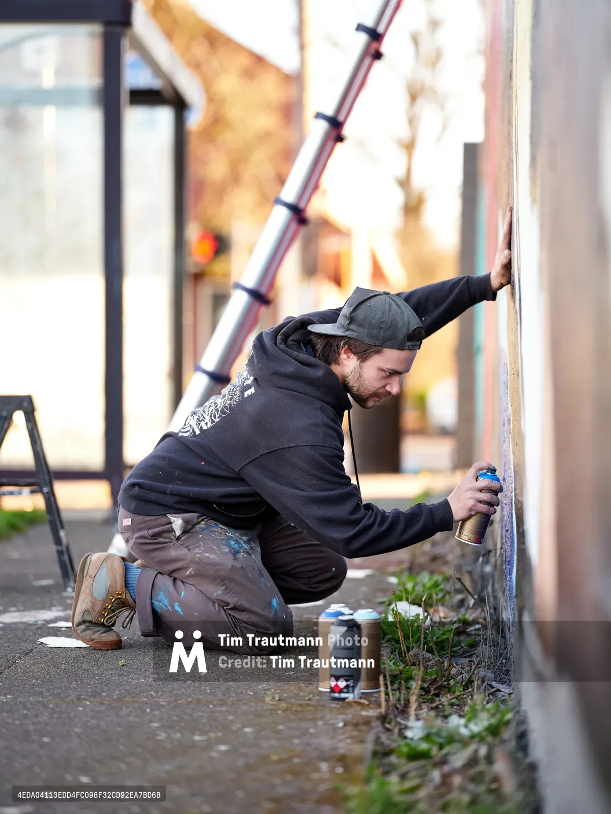 Travis Fields, known as campographic, kneels intently against a weathered concrete wall, spray can in hand as he works on his tribute mural to poet Amanda Gorman. The artist, wearing a dark hoodie and backwards cap, is captured mid-creation in Portland's Alberta Arts District, with paint-stained clothes telling the story of his artistic process. Scattered spray cans and wild grass growing from sidewalk cracks frame the intimate moment of urban art in progress.