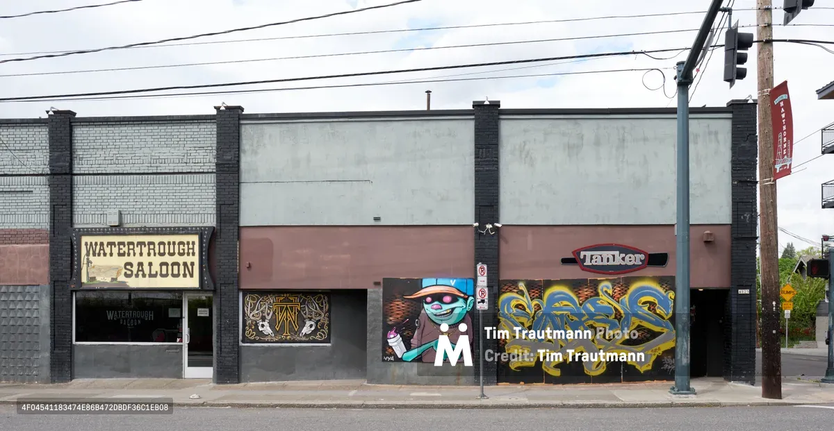 A weathered commercial strip along Southeast Hawthorne Boulevard in Portland reveals the stark reality of economic displacement. The Watertrough Saloon stands shuttered behind painted plywood, its rustic signage a ghost of livelier times, while vibrant street murals—including a cartoonish character in sunglasses and flowing yellow graffiti—create an ironic contrast against the abandoned storefronts. Power lines slice across an overcast sky, framing this tableau of urban decay and artistic resilience in Portland's Richmond neighborhood.
