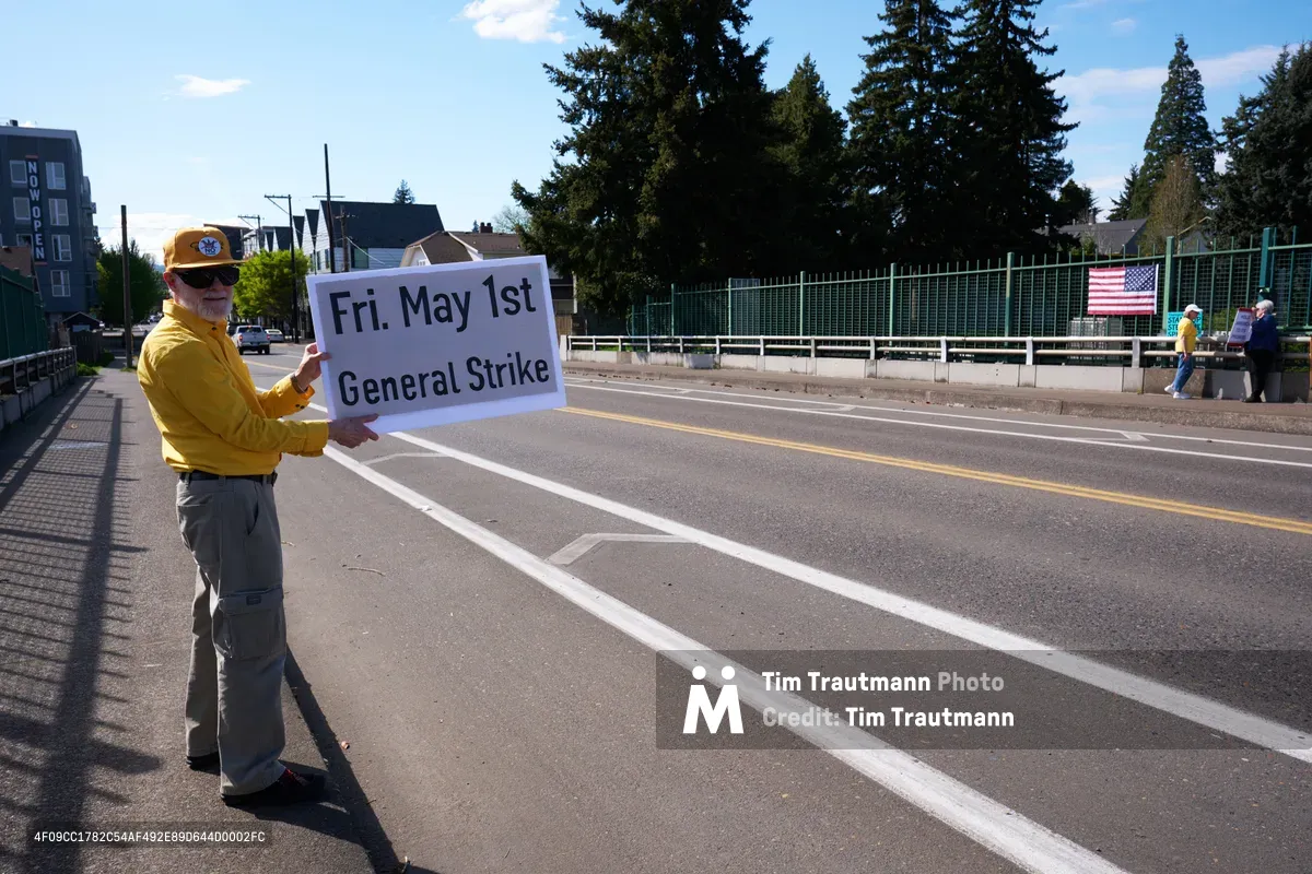 A determined activist in a bright yellow jacket and baseball cap stands on North Skidmore Street in Portland, holding a handmade sign announcing a May 1st General Strike. The bearded man positions himself against the stark urban backdrop of apartment buildings and evergreen trees under an expansive Pacific Northwest sky. His solitary but resolute stance embodies grassroots labor organizing in the quiet residential neighborhood of Humboldt.