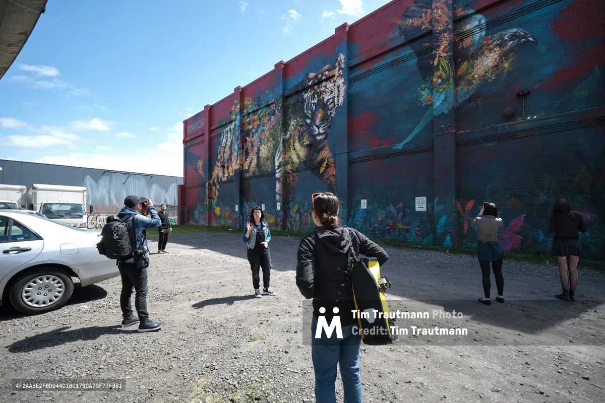A small group of visitors gathers on weathered asphalt before a monumental jaguar mural that dominates the industrial facade of a Central Eastside warehouse. Tour guide Tiffany Conklin gestures toward the vibrant street art while participants with cameras and backpacks maintain social distance under the crystalline Oregon sky. The scene captures the intersection of urban renewal and artistic expression in Portland's evolving industrial landscape.
